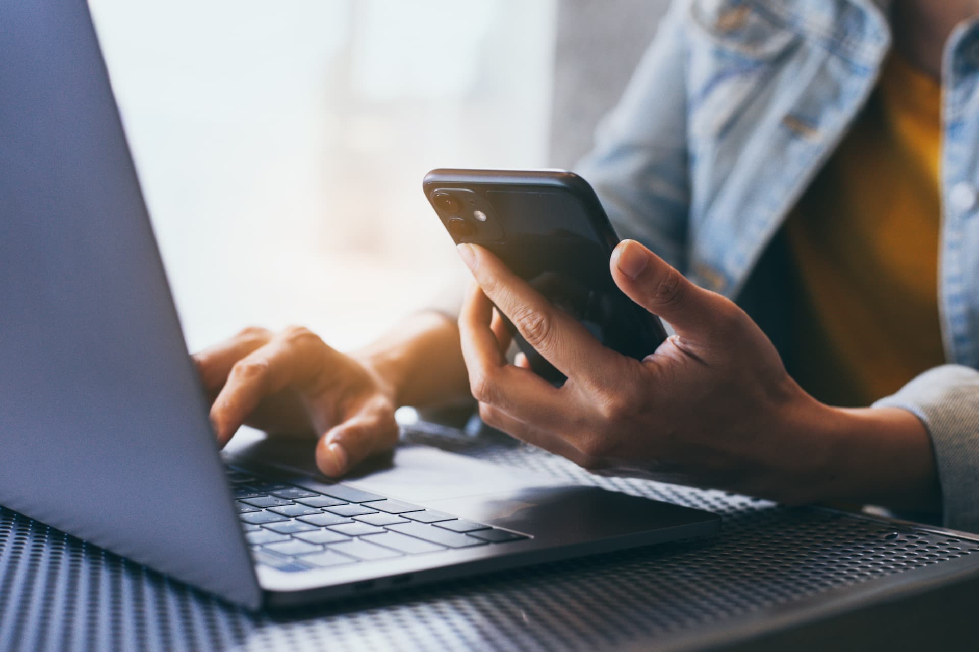 A person sitting at a desk using a laptop and looking at a phone in their hand